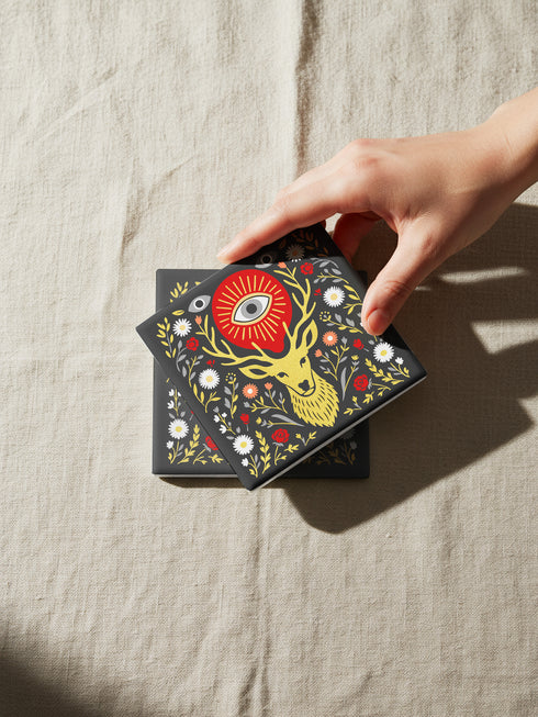 Square ceramic coaster, 4x4 inches, with cork backing. Charcoal tile showcases a golden stag head with antlers, surrounded by florals and an all-seeing eye in red and gold. Smooth printed surface; bold folk-art style. Witchy occult deer coaster for dark academia décor, bar cart, or living room. Square ceramic drink coaster with cork base, stag and eye design.